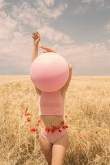 A woman wearing a pink top and matching bottoms, adorned with a garland of red poppies around her waist, holds a large pink balloon in front of her face. She stands in a field of golden wheat with a bright blue sky above.